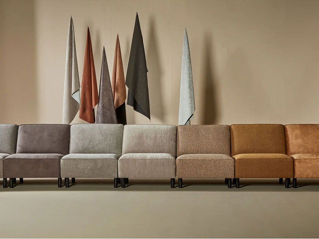 Eine Reihe verschiedener modularer Sofas in neutralen Tönen, hinter denen vor einer beigen Wand Stoffmuster hängen.